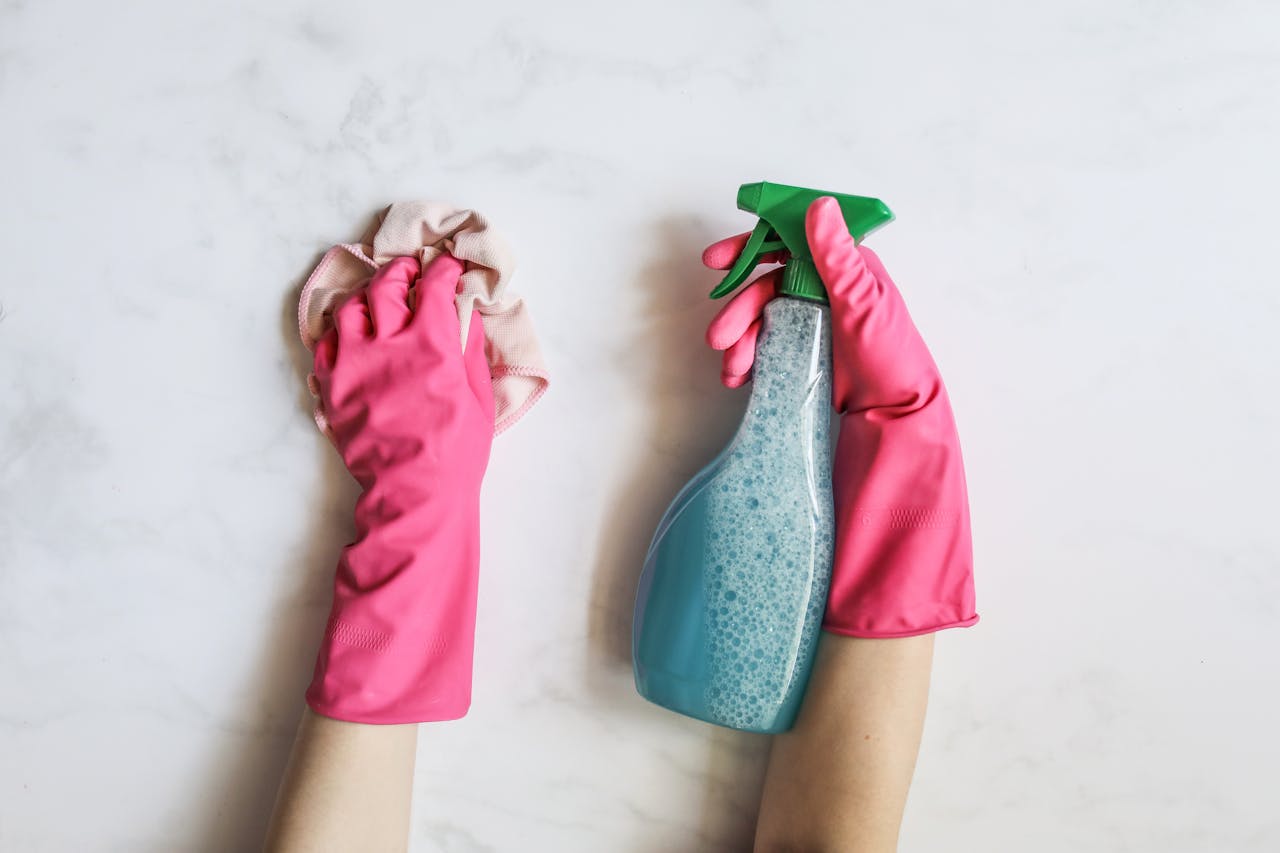 our-story A pair of hands in pink gloves are cleaning with a spray bottle on a white background.