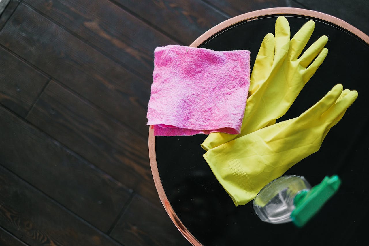 gallery-01 Yellow gloves and pink cloth on a table with cleaner bottle, viewed from above.
