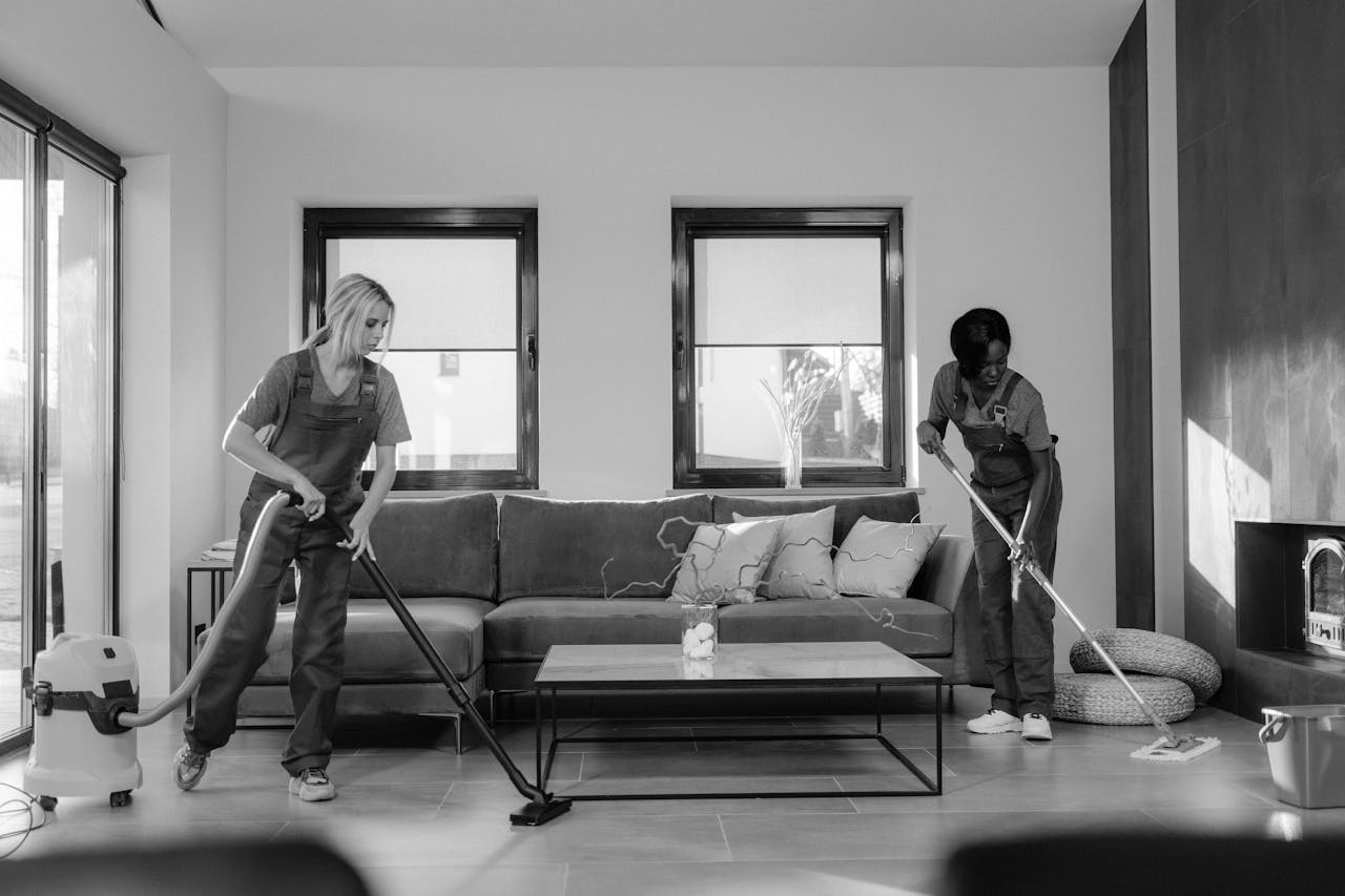 home-img Two women in work clothes cleaning a modern living room using a vacuum and mop.